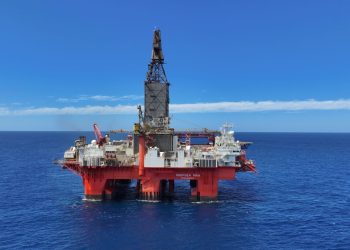 Offshore oil rig/platform floating on calm blue ocean under a clear sky, with a tall drilling tower.