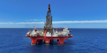 Offshore oil rig/platform floating on calm blue ocean under a clear sky, with a tall drilling tower.