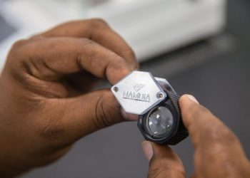 Close-up of hands holding a small handheld gem inspection tool with a hexagonal metal plate and a lens.