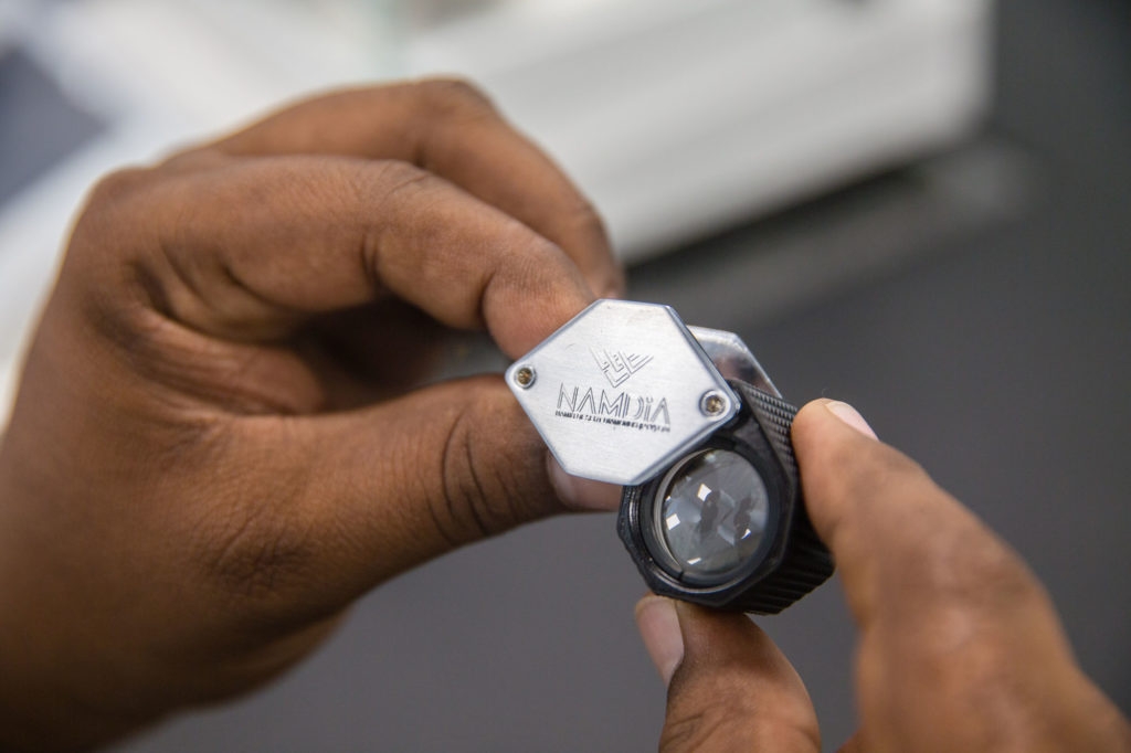 NAMDIA | Mining and Energy Namibia | Namibia’s Leading Mining & Energy News Close-up of hands holding a small handheld gem inspection tool with a hexagonal metal plate and a lens.