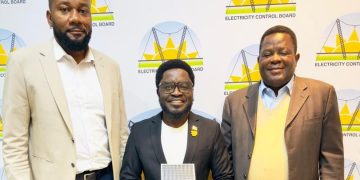Three men in suits pose for a photo at an Electricity Control Board event, the center man holding a yellow trophy in front of branded backdrop.