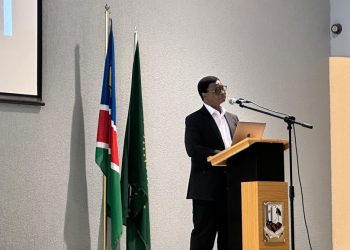 A man in a dark suit speaks at a wooden podium with a laptop, microphone, and two flag poles behind him in a conference room.