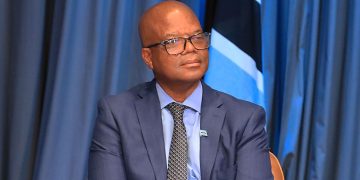 Man in a navy suit and glasses sits on stage with a blue curtain backdrop.
