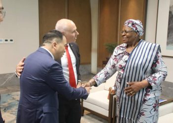 Two men in suits greet an African woman in a patterned outfit with a warm handshake in a formal lobby setting, smiling faces evident.