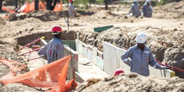 Construction workers wearing hard hats stand beside a dug trench with concrete forms on a sunny construction site.
