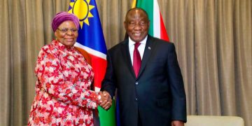Two officials shake hands in front of national flags during a formal meeting, with beige curtains in the background.