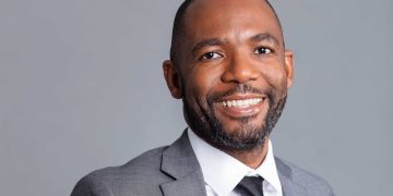 Professional headshot of a smiling man in a gray suit, white shirt, and black tie against a light gray background.