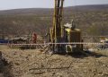 Oil/gas drilling rig in a rocky arid landscape with a worker in red near a wooden platform and hoses; site is cordoned off with tape and equipment nearby.