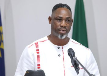 Hofni Iipinge | Mining and Energy Namibia | Namibia’s Leading Mining & Energy News Man speaking at a podium with a microphone, wearing a white traditional outfit, with flags in the background.