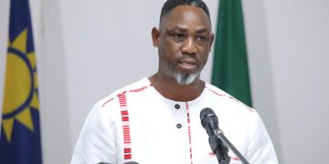 Man speaking at a podium with a microphone, wearing a white traditional outfit, with flags in the background.