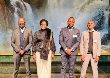 Four professionally dressed people stand in a row, smiling, in front of a waterfall backdrop.