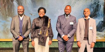 Four professionally dressed people stand in a row, smiling, in front of a waterfall backdrop.