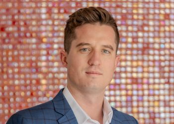Portrait of a man in a blue check suit jacket and white shirt, posed in front of a colorful mosaic backdrop.