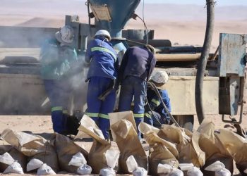 FRONTIER | Mining and Energy Namibia | Namibia’s Leading Mining & Energy News Industrial workers in blue uniforms and hard hats operate a concrete mixer in a desert setting, with bags of material laid out front.