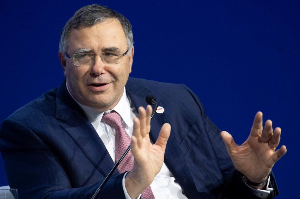 Man in a dark pinstripe suit and pink tie speaks into a microphone, hands raised in explanation against a solid blue background.