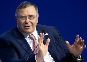 Patrick Pouyanné | Mining and Energy Namibia | Namibia’s Leading Mining & Energy News Man in a dark pinstripe suit and pink tie speaks into a microphone, hands raised in explanation against a solid blue background.