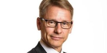 Professional headshot of a middle-aged man in a dark suit and red tie, wearing glasses, looking at the camera.