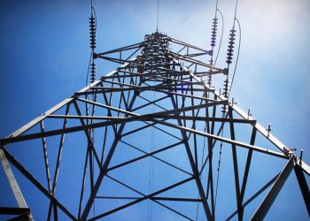 Low-angle view of a tall metal lattice transmission tower with insulators and power lines against a blue sky.