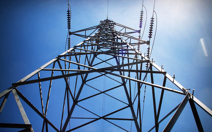 Low-angle view of a tall metal lattice transmission tower with insulators and power lines against a blue sky.