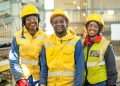Namdeb Employees in the Plant | Mining and Energy Namibia | Namibia’s Leading Mining & Energy News Three construction workers wearing yellow hard hats and high‑visibility gear smile inside an industrial facility.