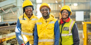 Three construction workers wearing yellow hard hats and high‑visibility gear smile inside an industrial facility.