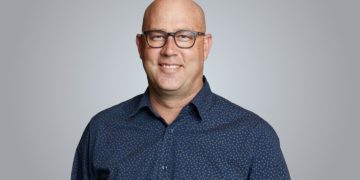 Smiling bald man wearing glasses and a navy patterned shirt, looking at the camera against a gray background.