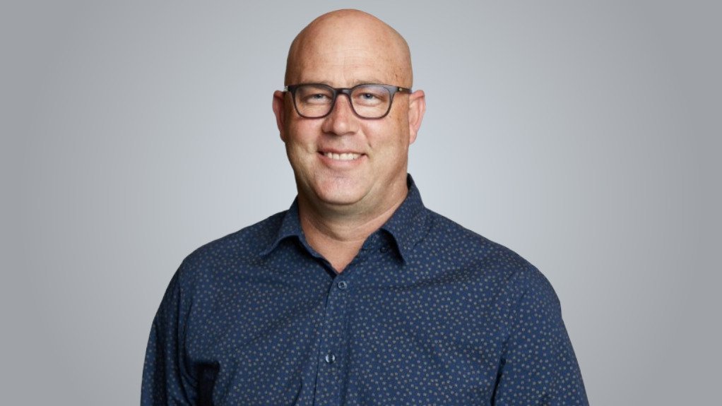 Smiling bald man wearing glasses and a navy patterned shirt, looking at the camera against a gray background.