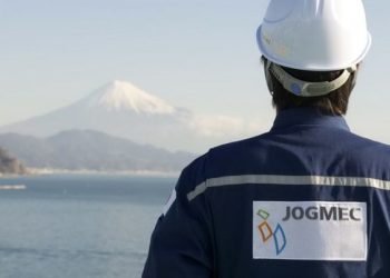 Back view of a worker in a white safety helmet and blue jacket with a JOGMEC patch, facing a tranquil coastal landscape with distant snow-capped mountains.