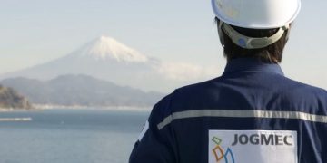 Back view of a worker in a white safety helmet and blue jacket with a JOGMEC patch, facing a tranquil coastal landscape with distant snow-capped mountains.