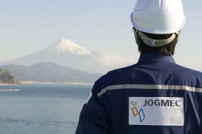 JOGMEC | Mining and Energy Namibia | Namibia’s Leading Mining & Energy News Back view of a worker in a white safety helmet and blue jacket with a JOGMEC patch, facing a tranquil coastal landscape with distant snow-capped mountains.