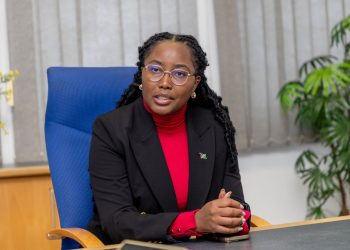 Emma mict 2 | Mining and Energy Namibia | Namibia’s Leading Mining & Energy News Professional woman seated at a desk in an office, wearing a red turtleneck and black blazer with a small lapel pin and gold earrings.