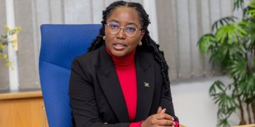 Emma mict 2 | Mining and Energy Namibia | Namibia’s Leading Mining & Energy News Professional woman seated at a desk in an office, wearing a red turtleneck and black blazer with a small lapel pin and gold earrings.