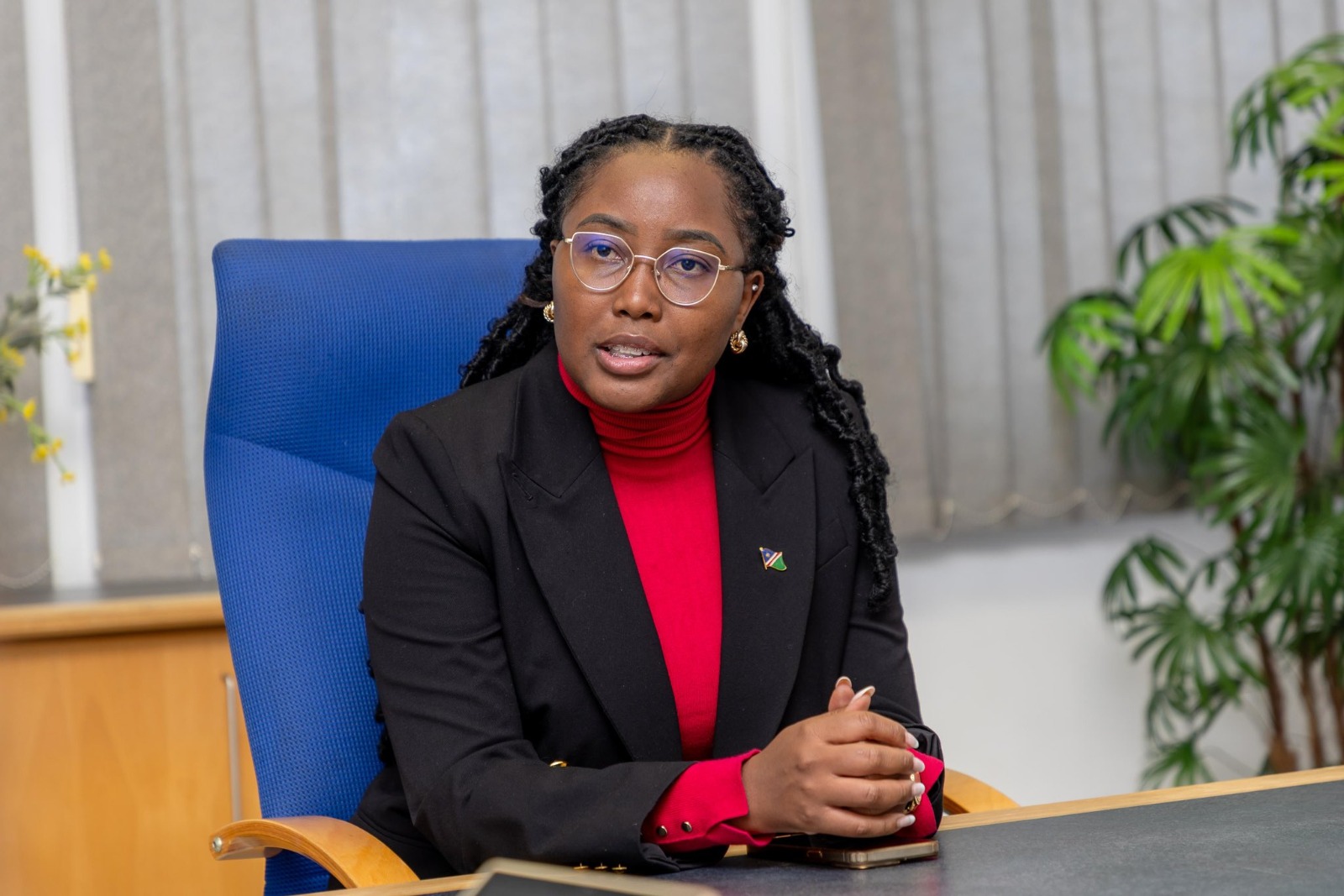 Professional woman seated at a desk in an office, wearing a red turtleneck and black blazer with a small lapel pin and gold earrings.