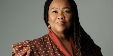 Portrait of a confident woman with long braids, wearing an orange-brown patterned top, looking at the camera.