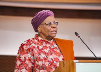 Older woman with a purple head wrap and floral dress speaks at a wooden podium with a microphone.