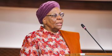 WhatsApp Image 2026-08-05 at 10539 PM | Mining and Energy Namibia | Namibia’s Leading Mining & Energy News Older woman with a purple head wrap and floral dress speaks at a wooden podium with a microphone.