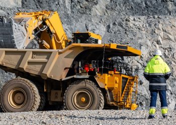 Two construction workers in high-visibility gear stand near a large yellow dump truck in a rocky quarry environment, inspecting the vehicle's cabin area.