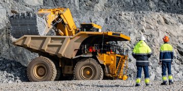 Two construction workers in high-visibility gear stand near a large yellow dump truck in a rocky quarry environment, inspecting the vehicle's cabin area.