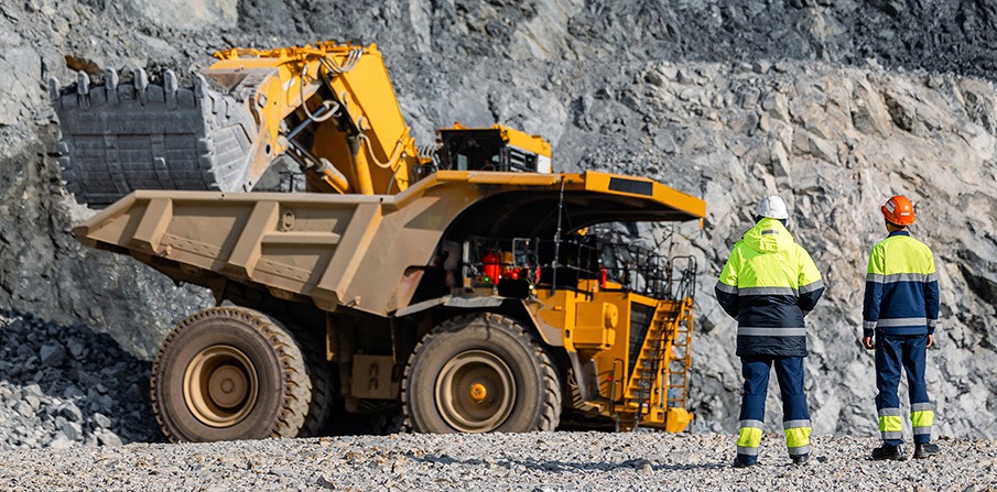 MINING 9 | Mining and Energy Namibia | Namibia’s Leading Mining & Energy News Two construction workers in high-visibility gear stand near a large yellow dump truck in a rocky quarry environment, inspecting the vehicle's cabin area.