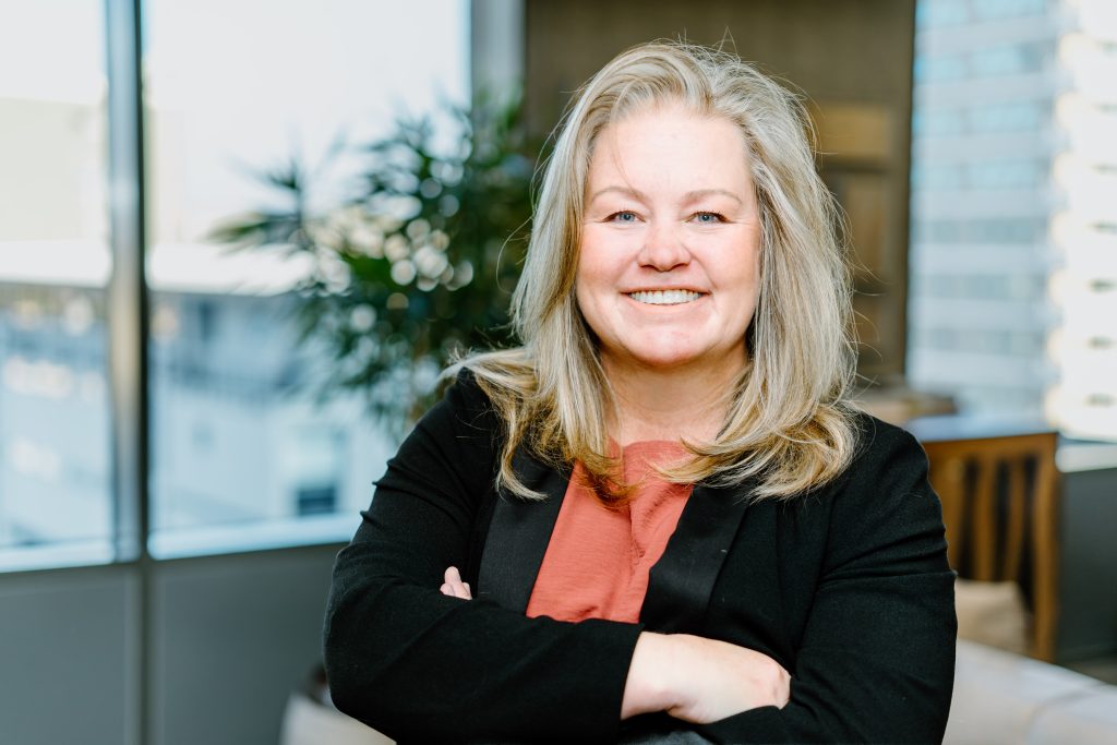 Angelique Peake | Mining and Energy Namibia | Namibia’s Leading Mining & Energy News Smiling woman with blonde hair wearing a coral blouse and black blazer, arms crossed in a bright office.