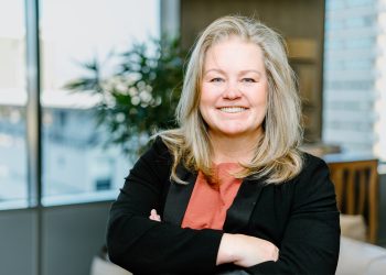 Angelique Peake | Mining and Energy Namibia | Namibia’s Leading Mining & Energy News Smiling woman with blonde hair wearing a coral blouse and black blazer, arms crossed in a bright office.