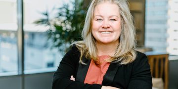 Angelique Peake | Mining and Energy Namibia | Namibia’s Leading Mining & Energy News Smiling woman with blonde hair wearing a coral blouse and black blazer, arms crossed in a bright office.