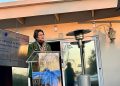 WhatsApp Image 2026-08-07 at 113449 AM | Mining and Energy Namibia | Namibia’s Leading Mining & Energy News Woman in a green blazer speaks at a clear podium on an outdoor terrace, with a screen showing a Finland mining sector event to her left.