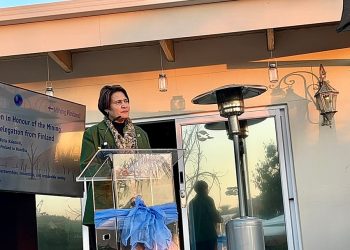 Woman in a green blazer speaks at a clear podium on an outdoor terrace, with a screen showing a Finland mining sector event to her left.