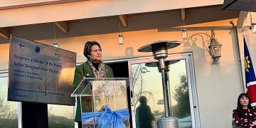 WhatsApp Image 2026-08-07 at 113449 AM | Mining and Energy Namibia | Namibia’s Leading Mining & Energy News Woman in a green blazer speaks at a clear podium on an outdoor terrace, with a screen showing a Finland mining sector event to her left.