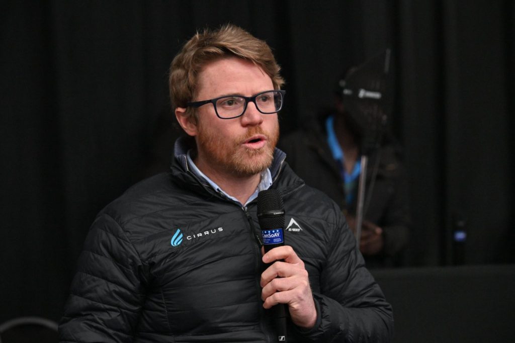 ROWLAND1 | Mining and Energy Namibia | Namibia’s Leading Mining & Energy News Man with glasses and a beard speaks into a handheld microphone on a dark stage, wearing a black quilted jacket with logos.