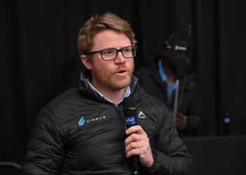 Man with glasses and a beard speaks into a handheld microphone on a dark stage, wearing a black quilted jacket with logos.