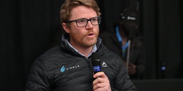 ROWLAND1 | Mining and Energy Namibia | Namibia’s Leading Mining & Energy News Man with glasses and a beard speaks into a handheld microphone on a dark stage, wearing a black quilted jacket with logos.