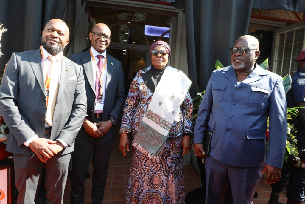 Four well-dressed adults posing outside a doorway: two men in suits, a woman in a patterned dress with a headwrap, and a man in a blue uniform; all wearing lanyards.