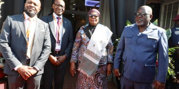 Four well-dressed adults posing outside a doorway: two men in suits, a woman in a patterned dress with a headwrap, and a man in a blue uniform; all wearing lanyards.